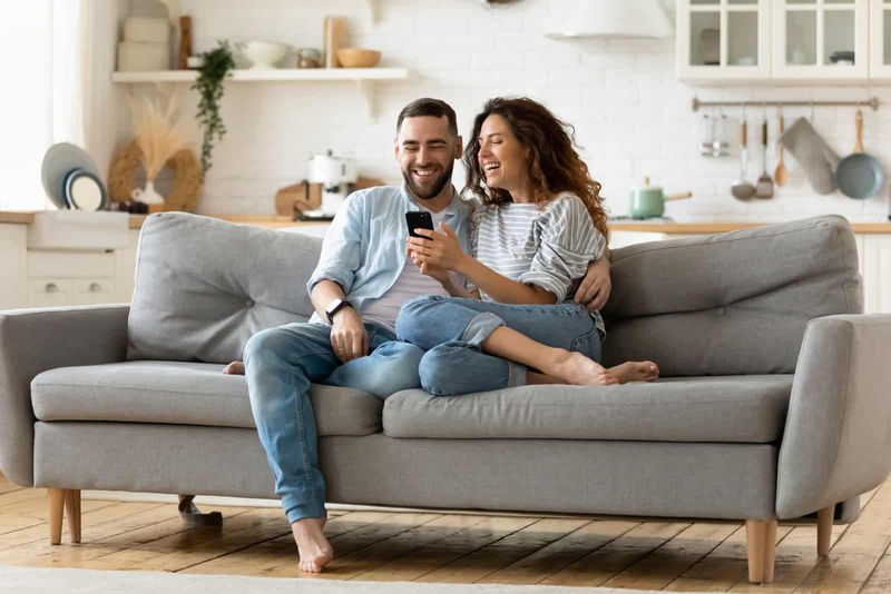 Ein lächelnder Mann und eine lächelnde Frau sitzen auf der Couch und schauen auf das Telefon