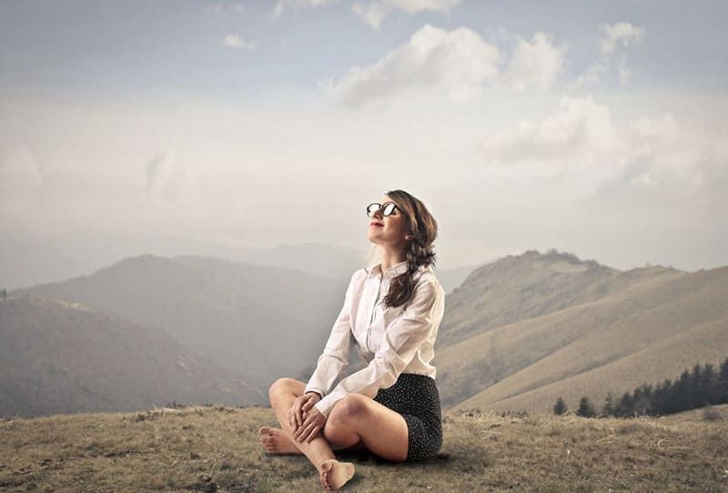 Friedliche, glückliche Frau, die barfuß auf dem Boden auf dem Gipfel des Berges sitzt und ein lässiges Outfit mit Brille trägt