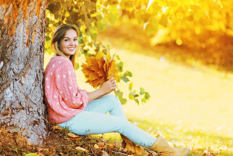 Fröhliche lächelnde Frau mit gelben Ahornblättern, die am sonnigen Herbsttag unter Baum sitzt