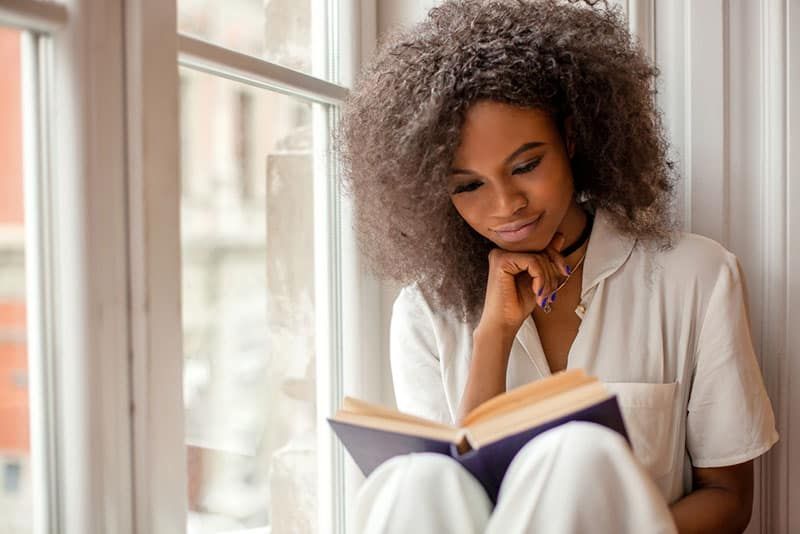 Hübsches afroamerikanisches Mädchen mit dem Lesen eines Buches auf der Fensterbank.