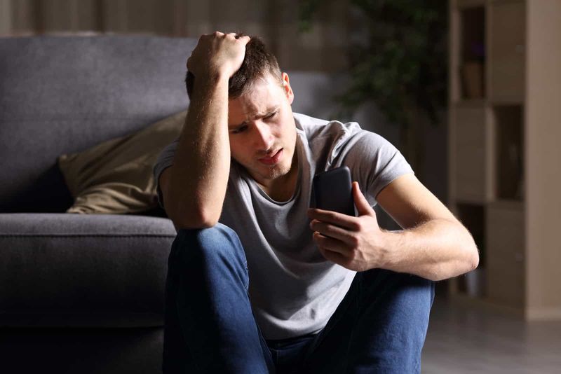Trauriger junger Mann, der auf dem Boden sitzt und auf sein Mobiltelefon schaut