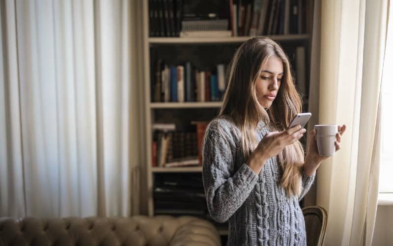 Frau im grauen Pullover, die eine weiße Keramiktasse hält, während sie auf ihrem Telefon eine SMS schreibt