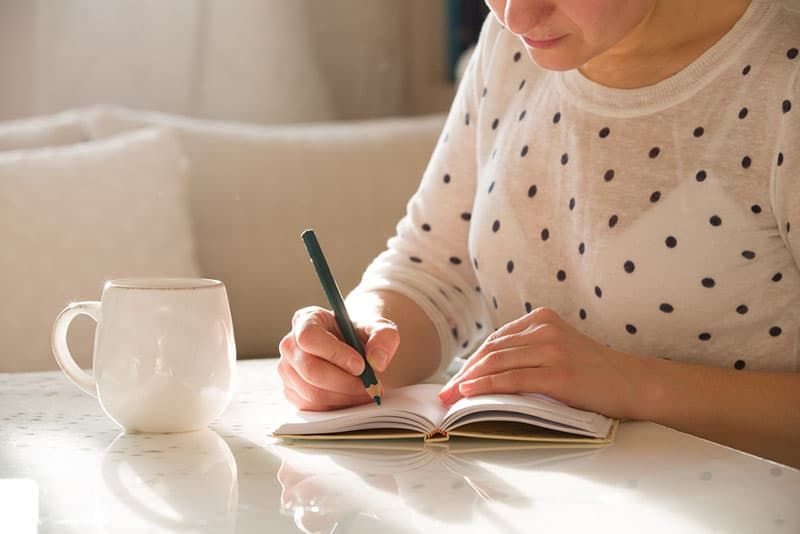 Frau sitzt am Tisch, schreibt in das Notizbuch und trinkt Kaffee in einem schönen, hellen Wohnambiente. Zuhause arbeiten. Freiberufler. Ideen aufschreiben. drinnen.
