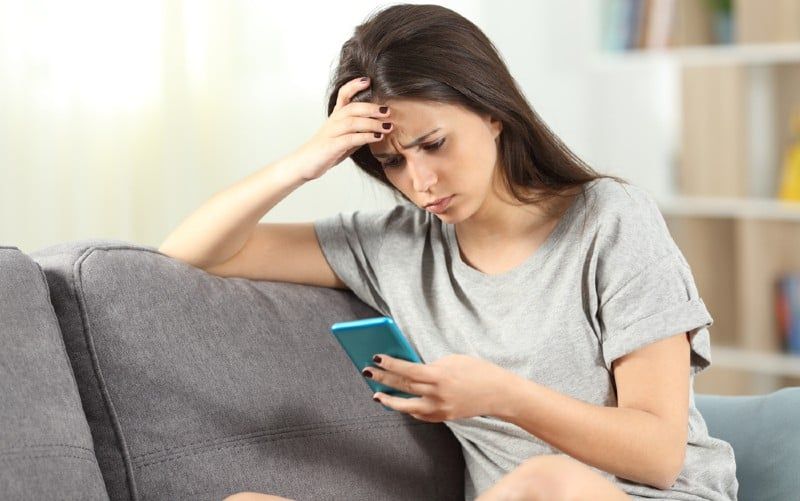 Trauriges Mädchen sitzt auf einer Couch und hält ein Smartphone in der Hand