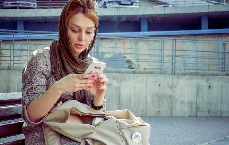 Frau mit Schal auf dem Kopf sitzt auf einer Bank und schreibt SMS auf ihr Handy