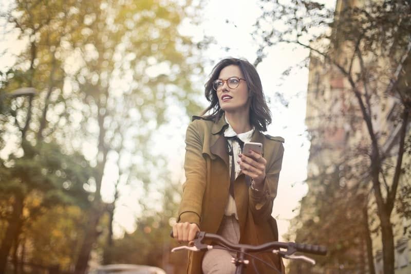 Frau, die Fahrrad fährt, während sie ein Smartphone mitten auf der Straße mit vielen Bäumen hält