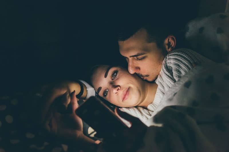 Mann küsst die Frau im Bett, während die Frau mit ihrem Smartphone beschäftigt ist