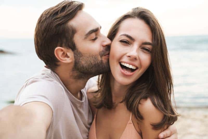 Mann küsst lächelnde Frau auf die Wange, während er ein Foto macht