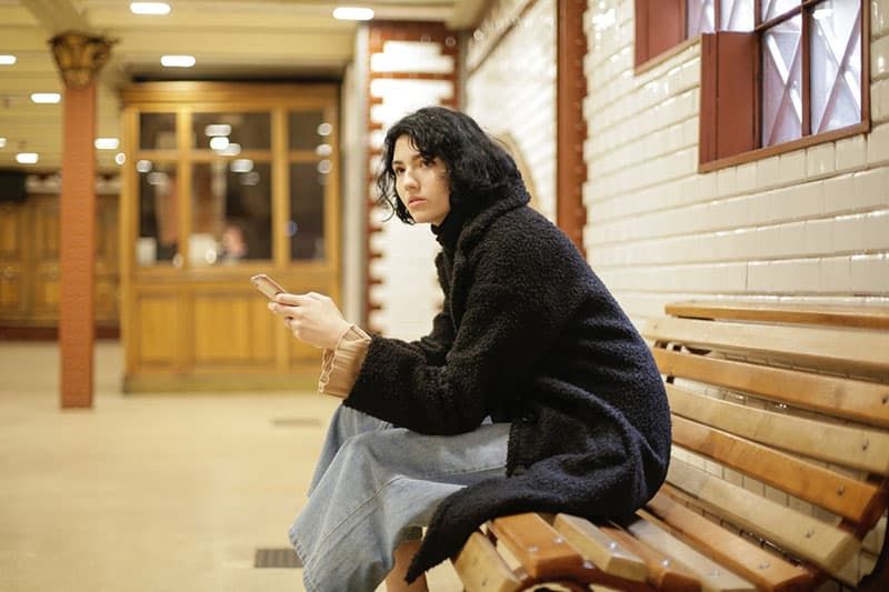 Frau sitzt auf einer Holzbank und wartet mit einem Mobiltelefon