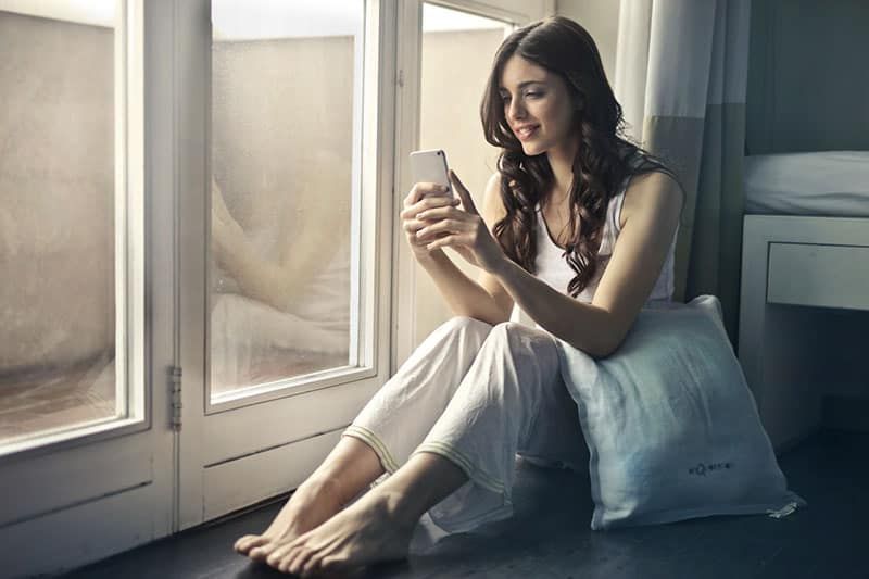Frau in Weiß hält Mobiltelefon und ein Kissen neben sich