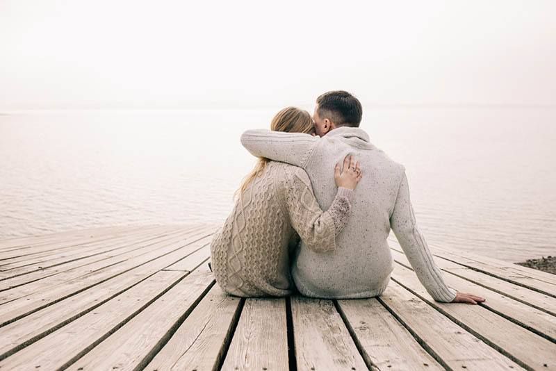 Rückansicht eines Paares, das auf einem Holzsteg sitzt und sich umarmt