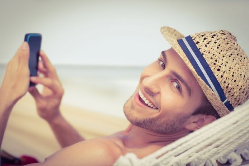 Mann entspannt sich in der Hängematte, schreibt SMS auf dem Smartphone und blickt lächelnd in die Kamera hinter ihm