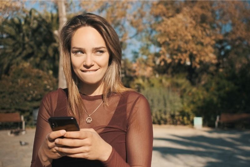 Flirtende blonde Frau, die draußen SMS am Telefon schreibt und schlau lächelt