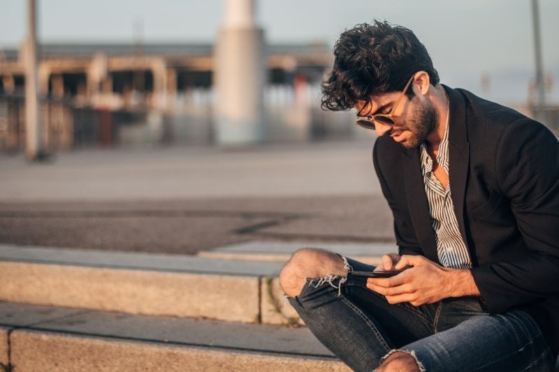 Gutaussehender Mann in schwarzer Jacke, der draußen mit dem Smartphone SMS schreibt