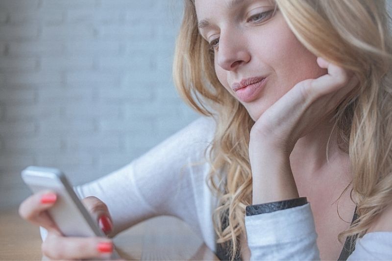 Junge nachdenkliche Frau schreibt SMS auf ihrem Smartphone, während sie mit einer Hand ihr Kinn stützt