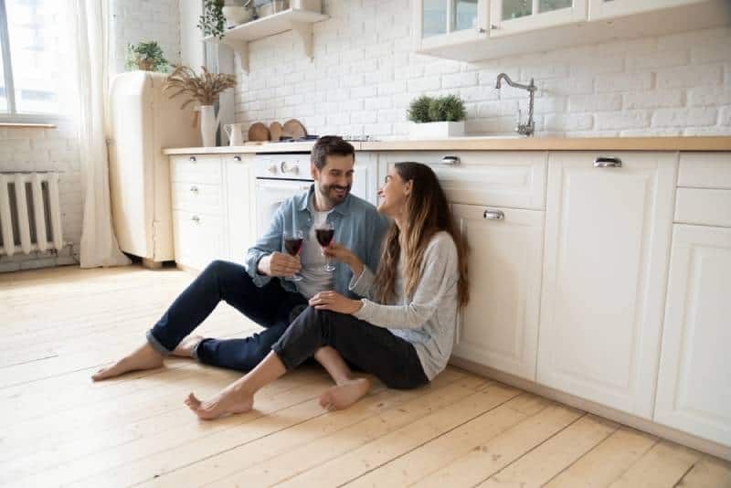romantisches paar sitzt auf dem küchenboden und trinkt wein, während sie sich lächelnd ansehen