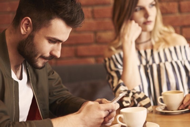 Ein Mann schreibt während eines Dates eine SMS in sein Mobiltelefon, während eine Frau gelangweilt aussieht und wegschaut
