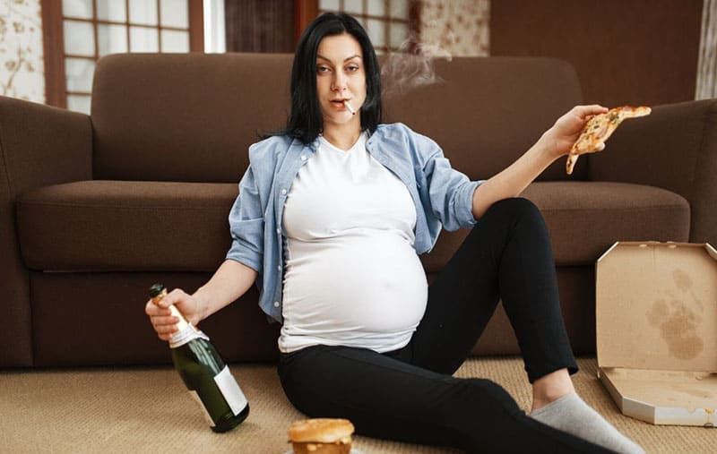Alkoholische schwangere Frau, die eine Flaschenpizza trägt, während sie auf dem Boden raucht