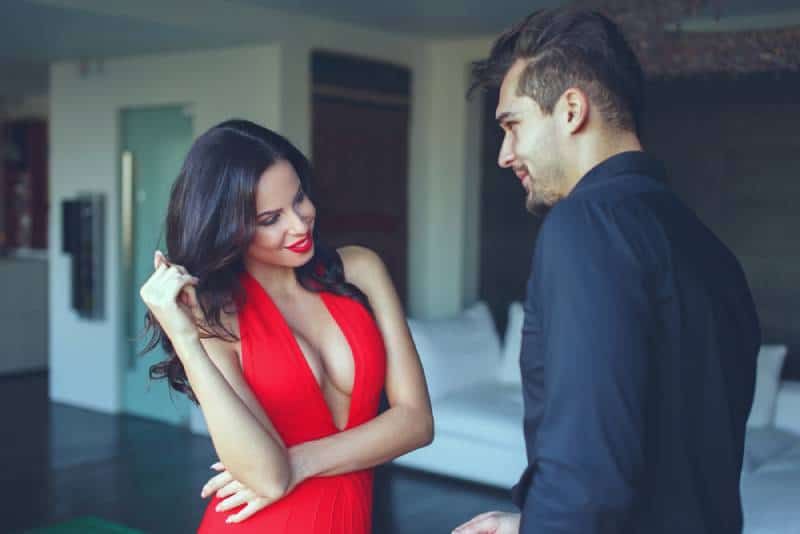 Frau in rotem Kleid und Lilie flirtet drinnen mit einem jungen Mann und spielt mit ihren Haaren