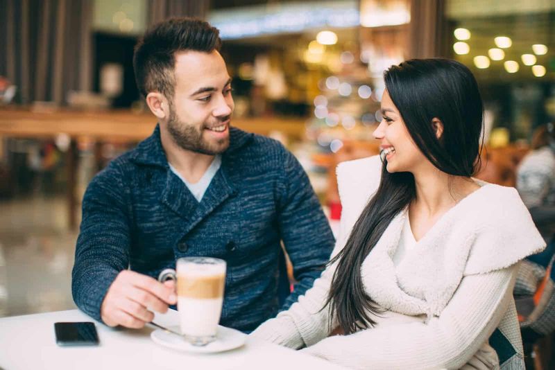 Eine lächelnde Frau mit langen schwarzen Haaren sitzt mit einem Mann in einem Café