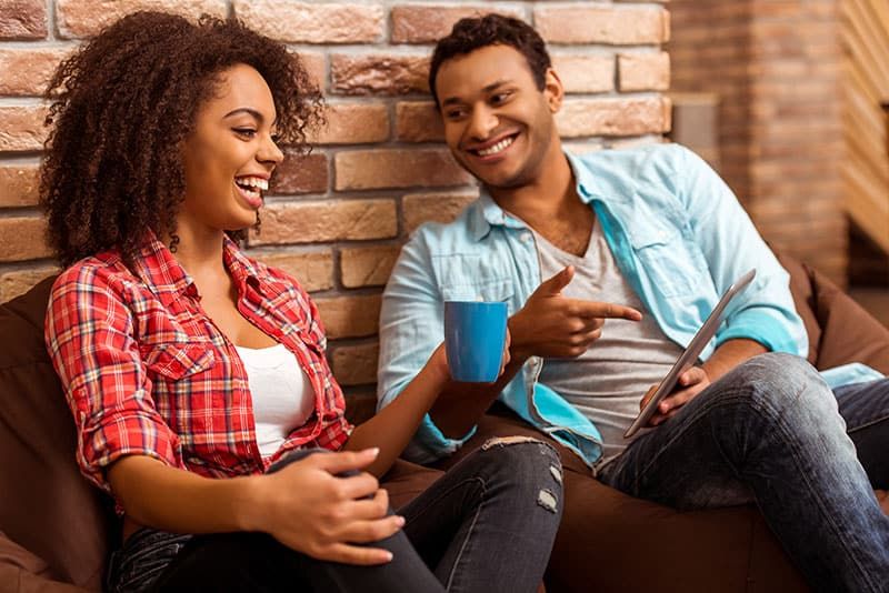 Attraktives afroamerikanisches Paar, das ein Tablet benutzt, eine Tasse hält und lacht, während es auf Sitzsäcken gegen eine Ziegelwand sitzt