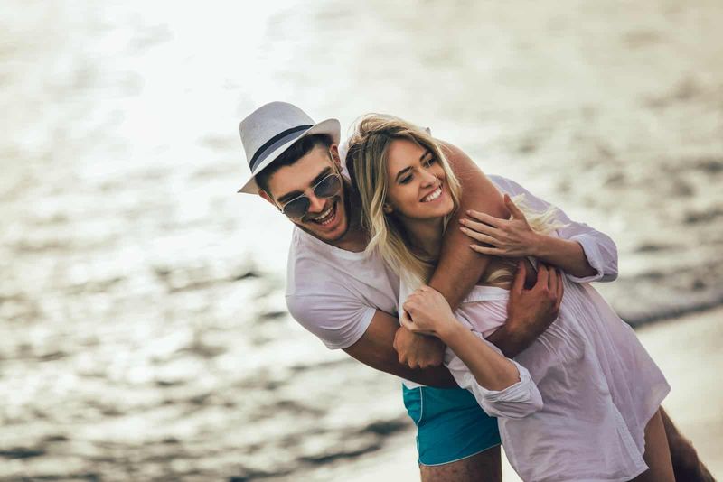 Ein lächelnder Mann mit Hut auf dem Kopf umarmt eine Frau am Strand und lacht
