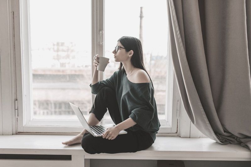 Junge Frau denkt darüber nach, Kaffee zu trinken, während sie mit einem Laptop auf dem Schoß am Fensterbrett sitzt