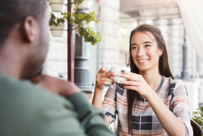 Eine lächelnde Frau sitzt mit einem schwarzen Mann in einem Café und trinkt Kaffee