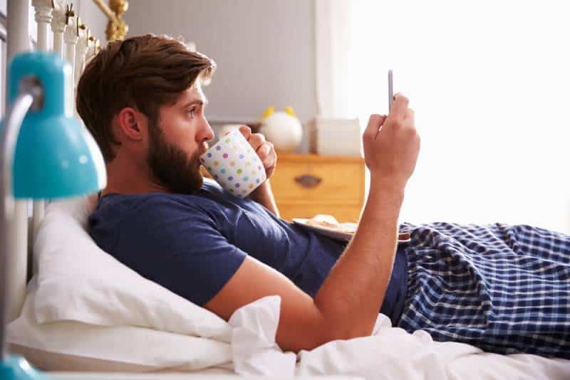 Mann im Schlafanzug liegt im Schlafzimmer und tippt auf seinem Telefon