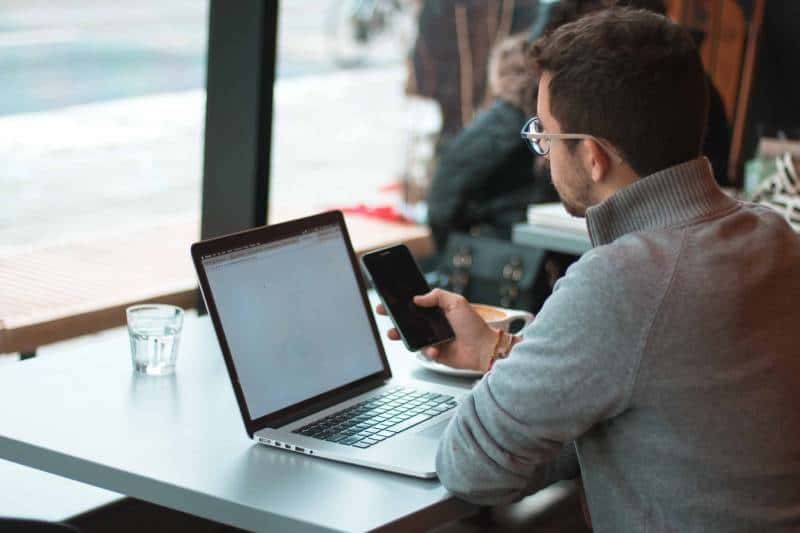 Mann sitzt am Tisch mit Laptop und Smartphone am Fenster