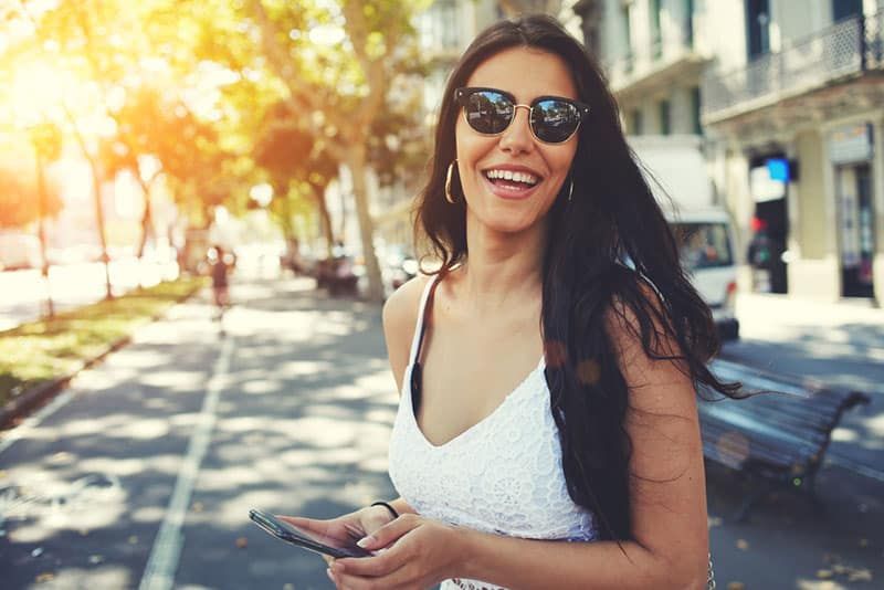 Fröhliche lateinamerikanische Frau mit Sommersonnenbrille, die hell lächelt, während sie das Smartphone hält, das an sonnigen Tagen im Freien in der Stadt spaziert, weibliche Person, die mobiles Stehen mit Kompositionskopierraum auf einer Seite verwendet