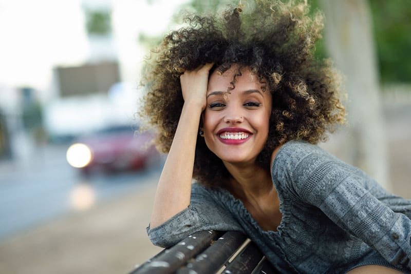 Junge gemischte Frau mit Afrofrisur, die im städtischen Hintergrund lächelt. Schwarzes Mädchen, das Freizeitkleidung trägt.