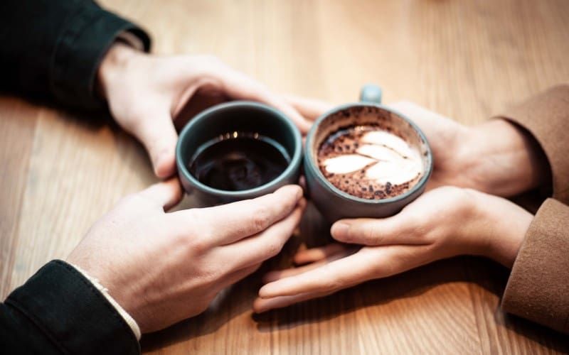 Die Hände zweier Personen, die Keramikbecher mit Kaffee auf einem braunen Tisch halten