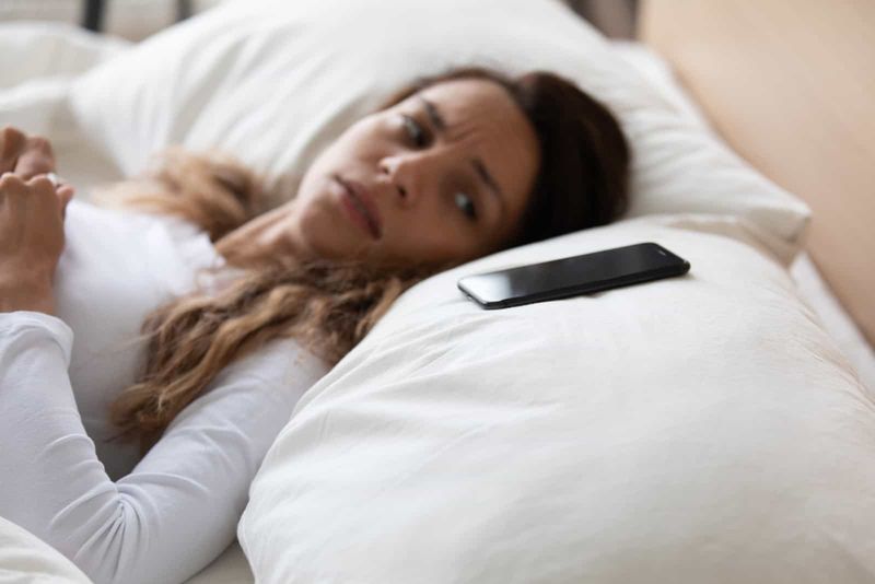 Müde Frau liegt im Bett und schaut auf ihr Telefon auf dem Kissen