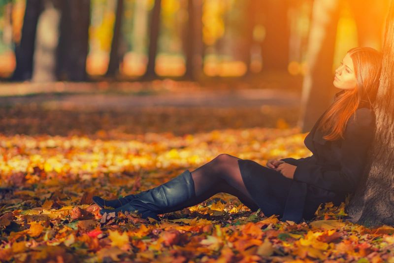 Frau in schwarzem Mantel und Lederstiefeln sitzt unter Baum im Herbstpark