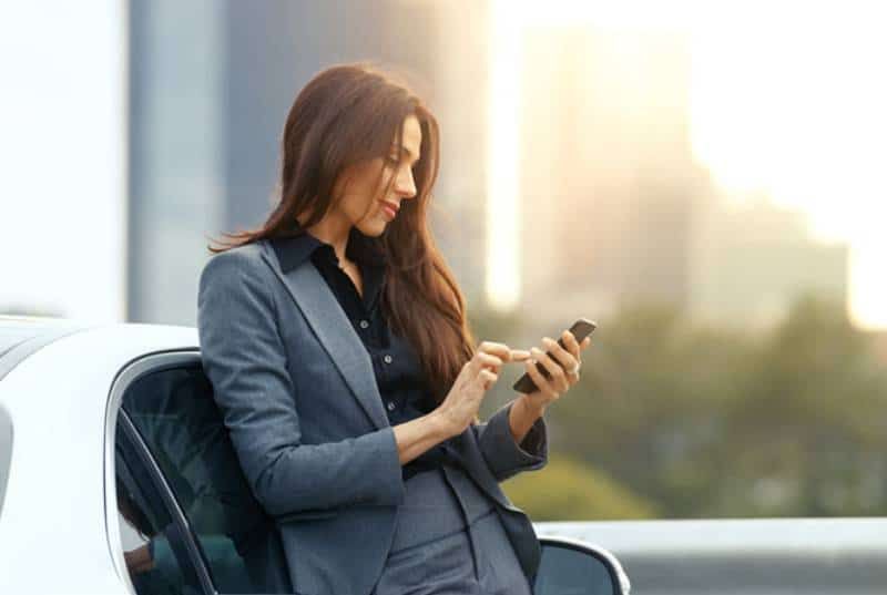 Lächelnde Geschäftsfrau, die auf seinem Telefon tippt, während sie sich draußen an das Auto lehnt