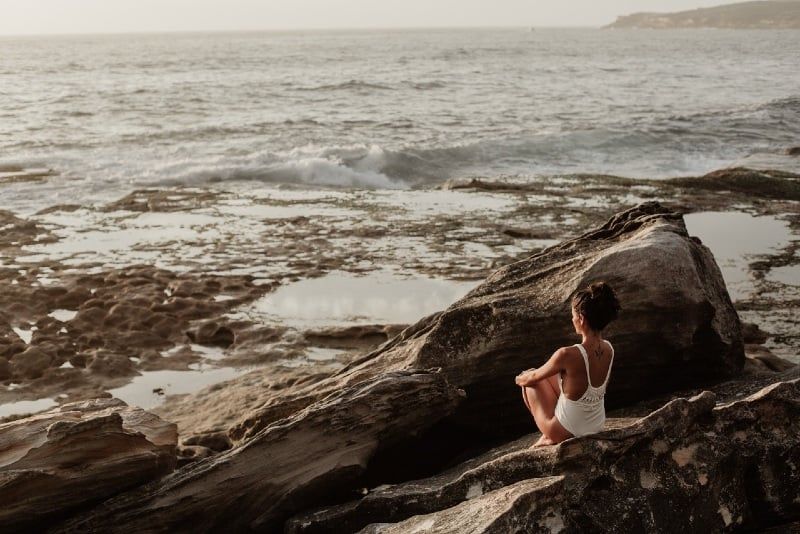 Frau im weißen Badeanzug sitzt auf Felsen und blickt aufs Meer