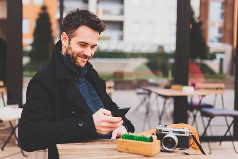 Lächelnder Mann im schwarzen Mantel, der im Straßencafé auf seinem Telefon tippt