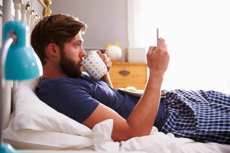 Ein Mann liegt auf dem Bett, trinkt Kaffee und drückt auf ein Telefon