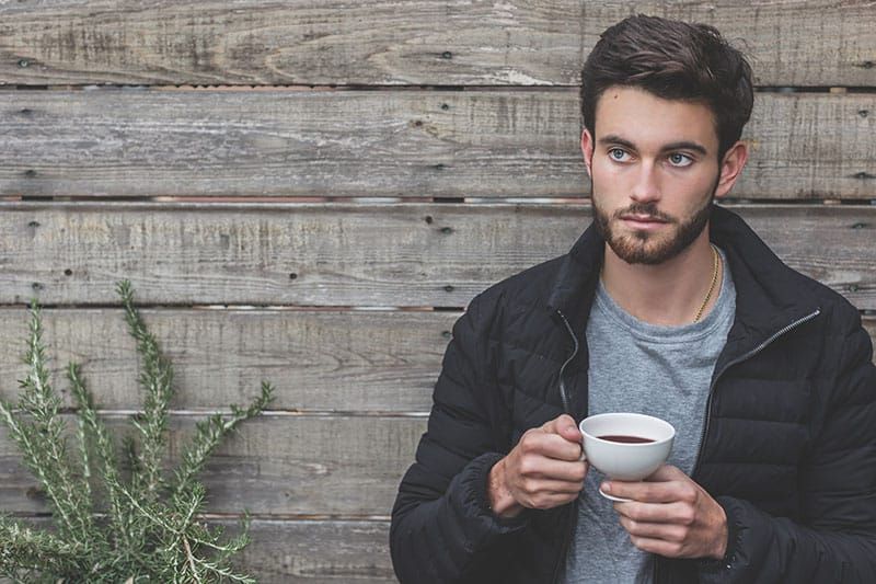 Mann hält eine Tasse Kaffee in der Hand, während er sich an eine Holzwand lehnt