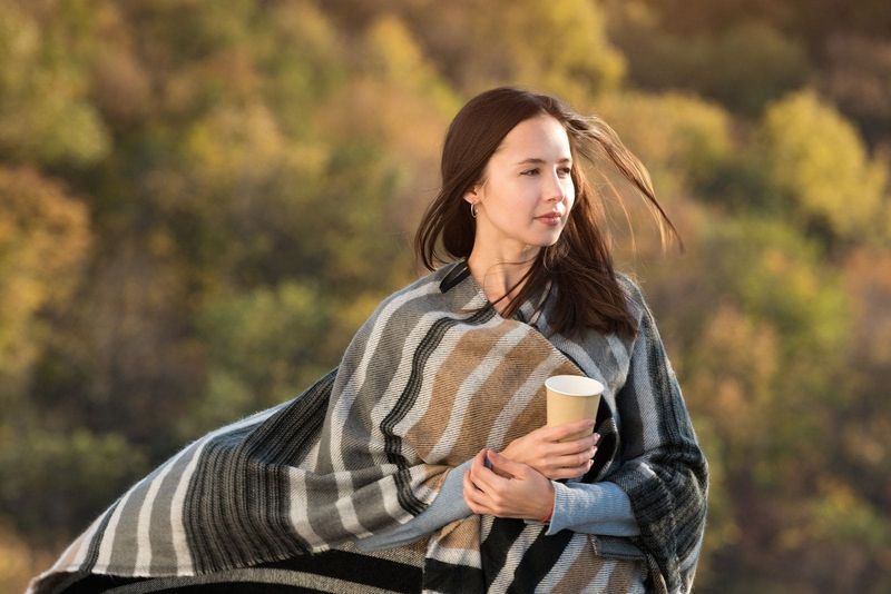Frau, eingewickelt in eine Decke, geht draußen und trägt Kaffee in einem Pappbecher