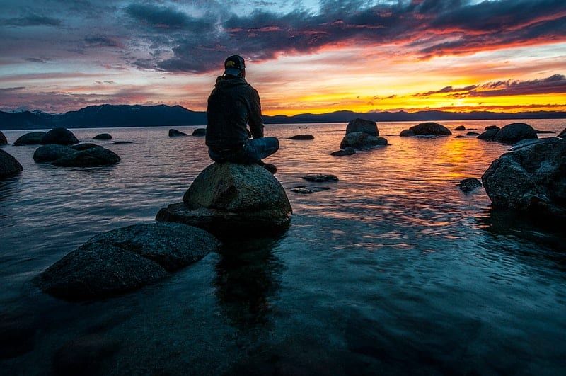 Person, die auf einem Felsen auf dem Wasser sitzt und einem Sonnenuntergang gegenübersteht