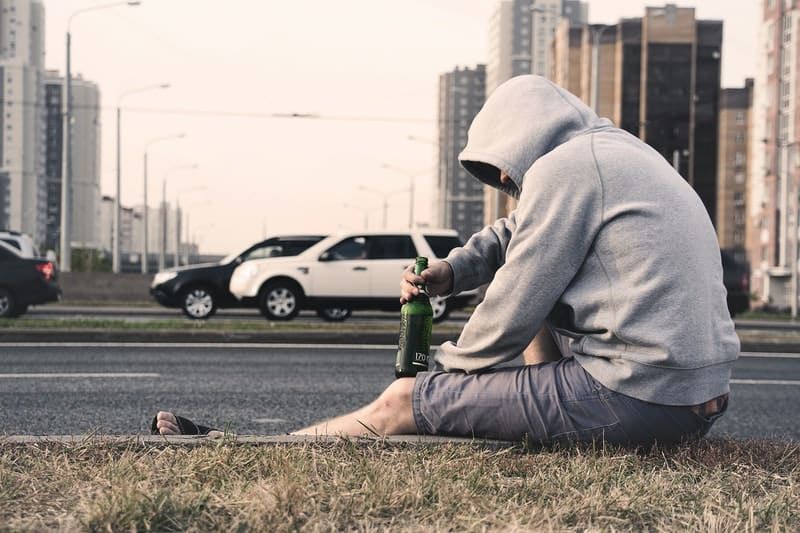 Alkoholiker auf der Straße, der Bier neben der Straße hält und ein Hemd mit Kapuze trägt