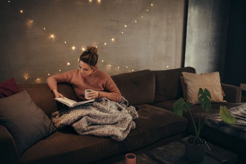 Junge Frau liest Buch und trinkt Kaffee auf dem Sofa im Hygge-Haus. Kaukasische Frauen, die sich im gemütlichen Wohnzimmer entspannen und ein Buch lesen.
