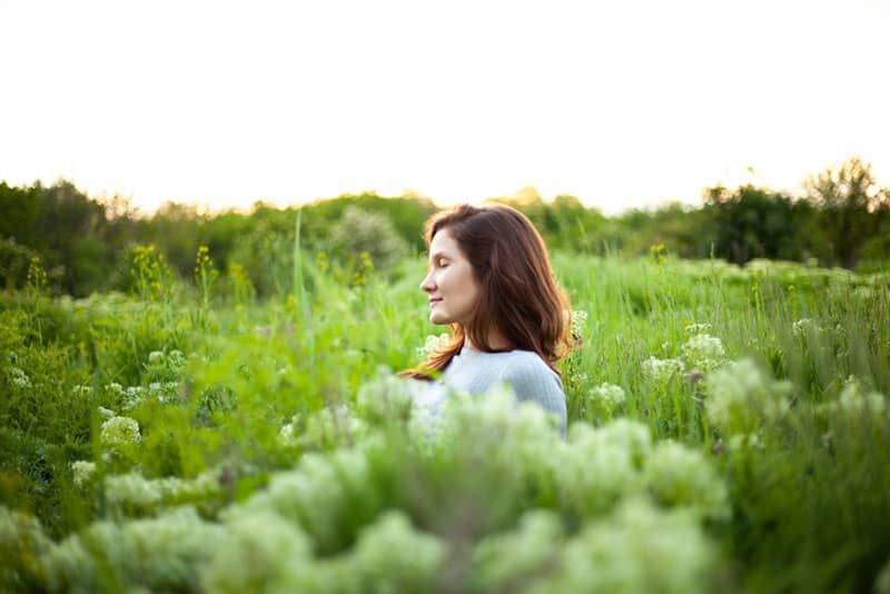 Schöne und attraktive Frau mit roten Haaren sitzt seitlich auf einem Feld und bewundert, genießt den Sonnenuntergang in den Bergen, Feld, Wiese