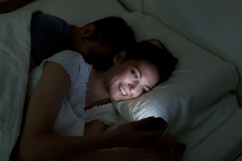 Eine lächelnde Frau im Bett mit einem schlafenden Mann benutzt ein Mobiltelefon