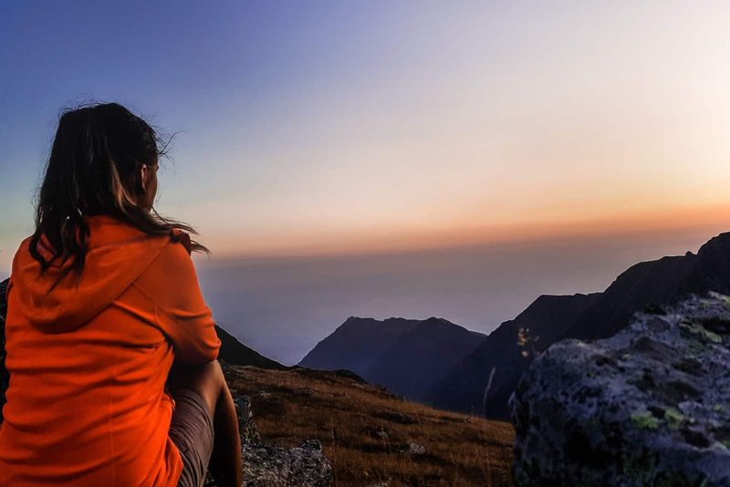 Frau in orangefarbener Jacke sitzt auf einem Felsen und blickt auf den Berg