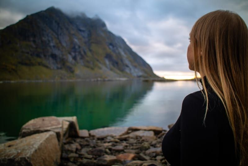 Frau in schwarzer Jacke sitzt auf einem Felsen und blickt auf den See