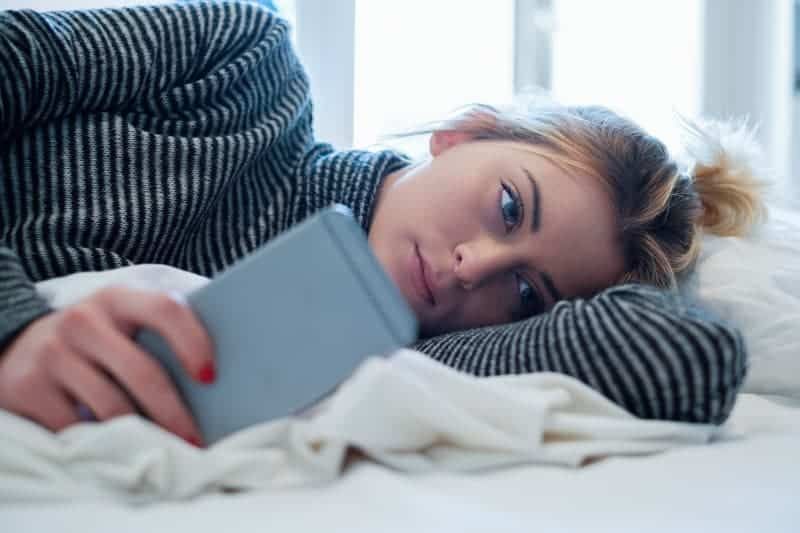 Nachdenkliche Frau, die auf dem Bett liegt und auf ihrem Telefon tippt