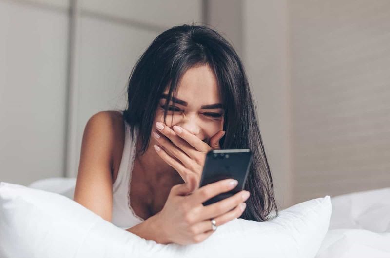 Mädchen lacht und starrt auf ihr Telefon, während sie auf dem Bett liegt